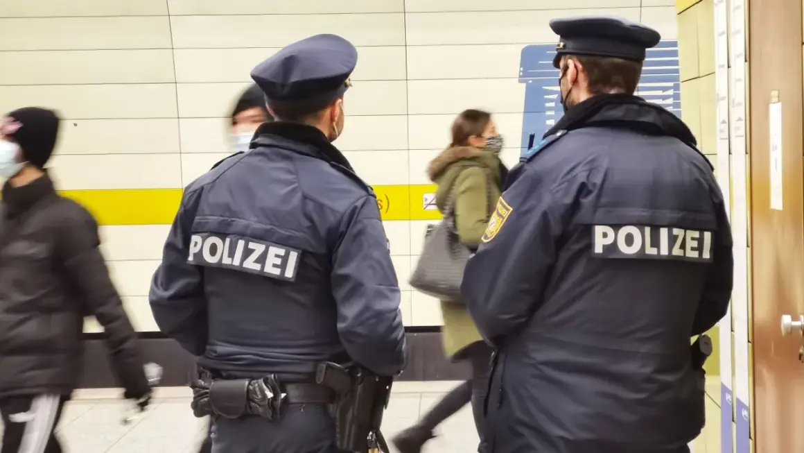 Zwei Polizisten stehen in einer U-Bahn-Station. Im Hintergrund sind 53 Personen zu sehen, die sich bewegen.