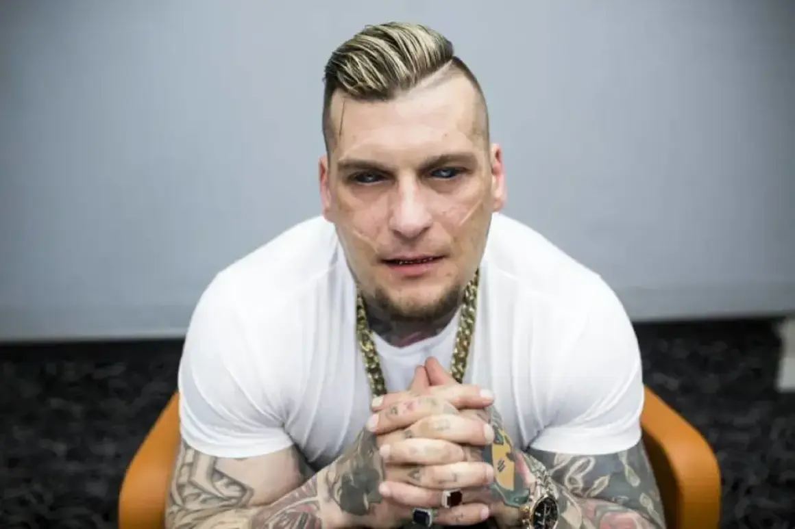 Popek, raper i zawodnik MMA, w białej koszulce i złotym łańcuchu, z tatuażami na rękach i twarzy.