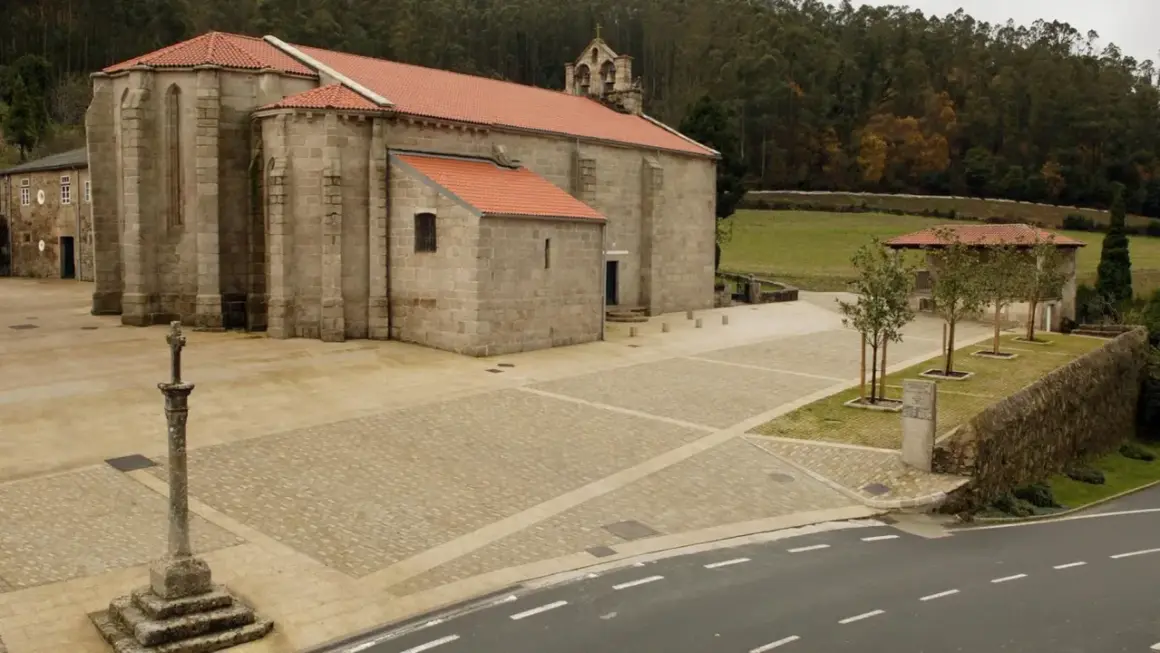 El monasterio de Soandres, con su piedra gris y tejados rojos, se alza majestuoso ante un bosque frondoso. Un cruceiro de piedra vigila la plaza empedrada.
