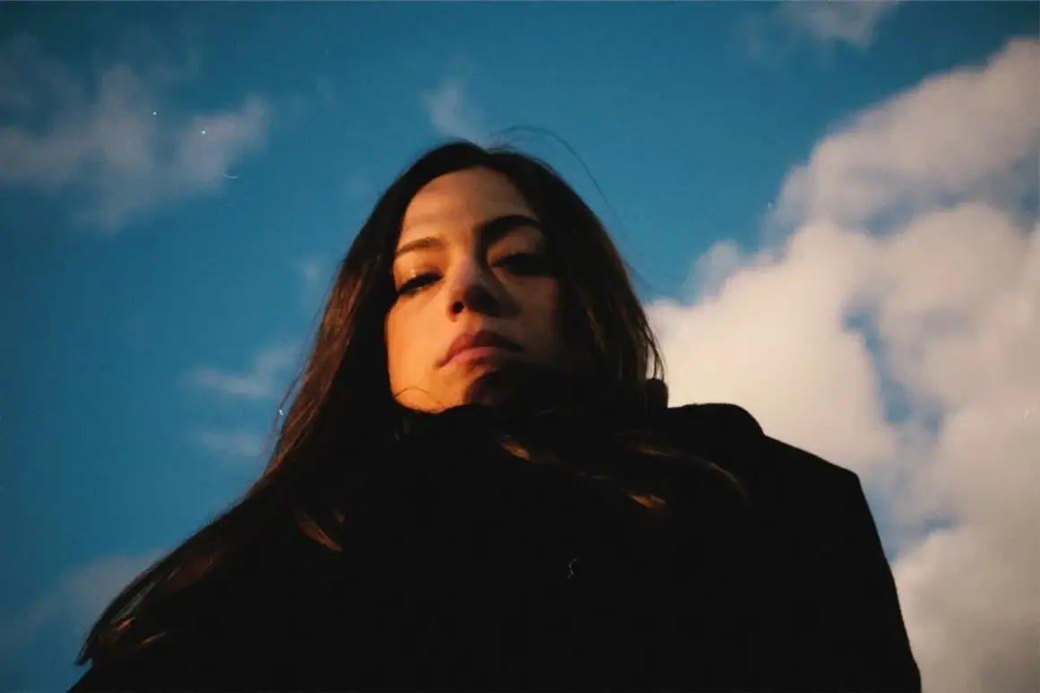 Daniela Pes spira sotto un cielo azzurro e nuvoloso, con uno sguardo intenso e capelli scuri che incorniciano il viso.