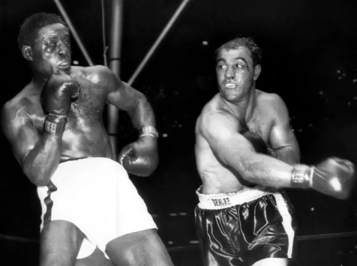 Two boxers, one with white shorts, the other with black, are in the ring. The boxer on the right, with a cut above his eye, is Ezra Charles.