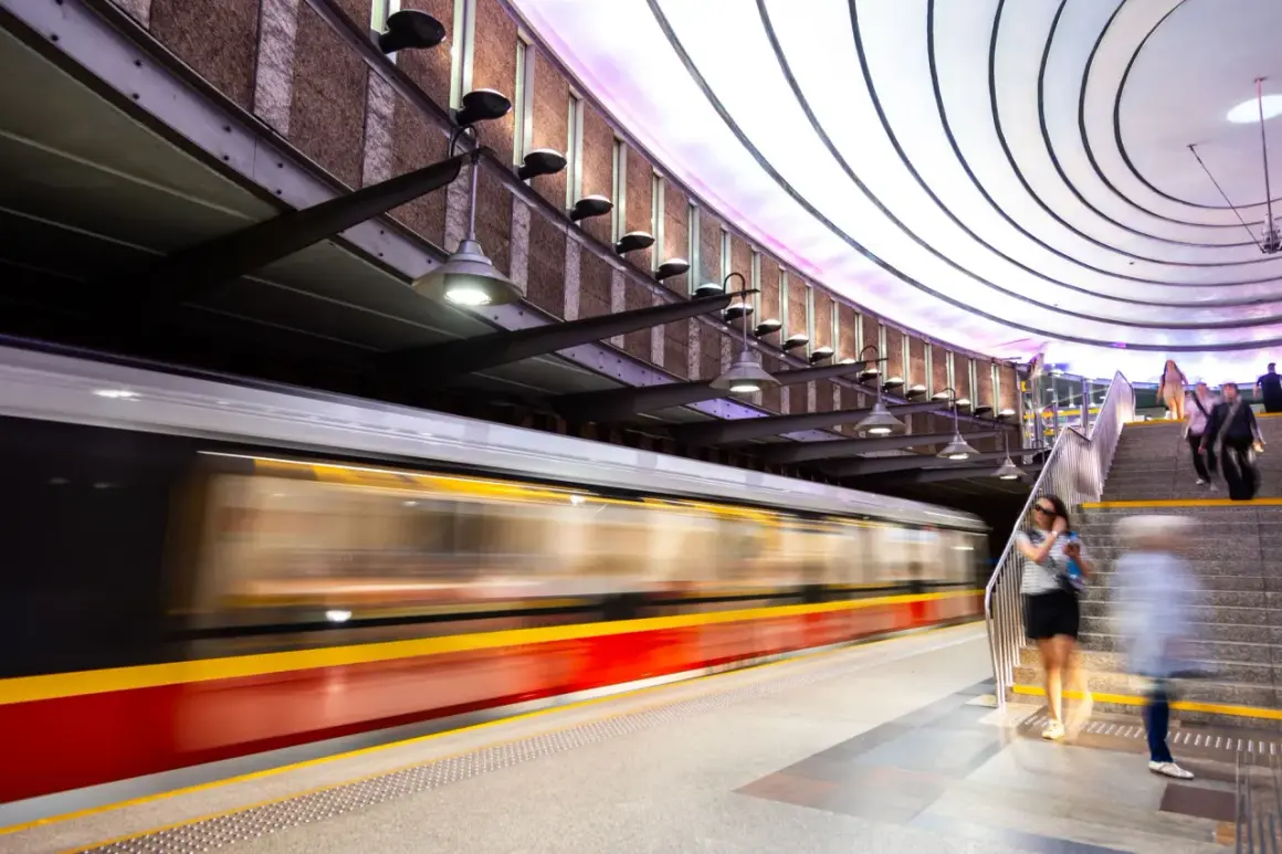 Nowoczesna stacja metra, do której jeździ metro. Pociąg mija peron, a ludzie schodzą po schodach.