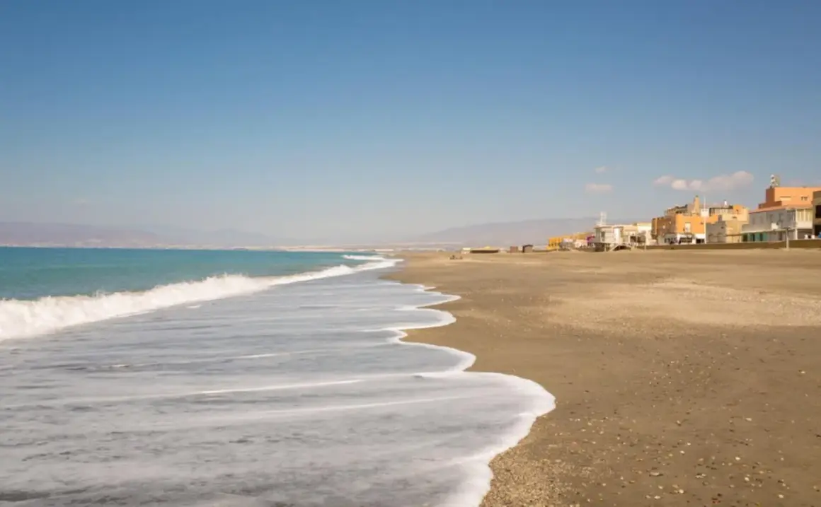 Olas suaves acarician la arena dorada de la playa de Cabo de Gata. Al fondo, un pueblo costero se alza bajo un cielo azul.