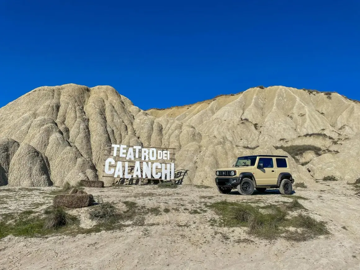 Un fuoristrada beige parcheggiato davanti al **teatro dei calanchi**, un paesaggio lunare con montagne erose e un cartello in legno.