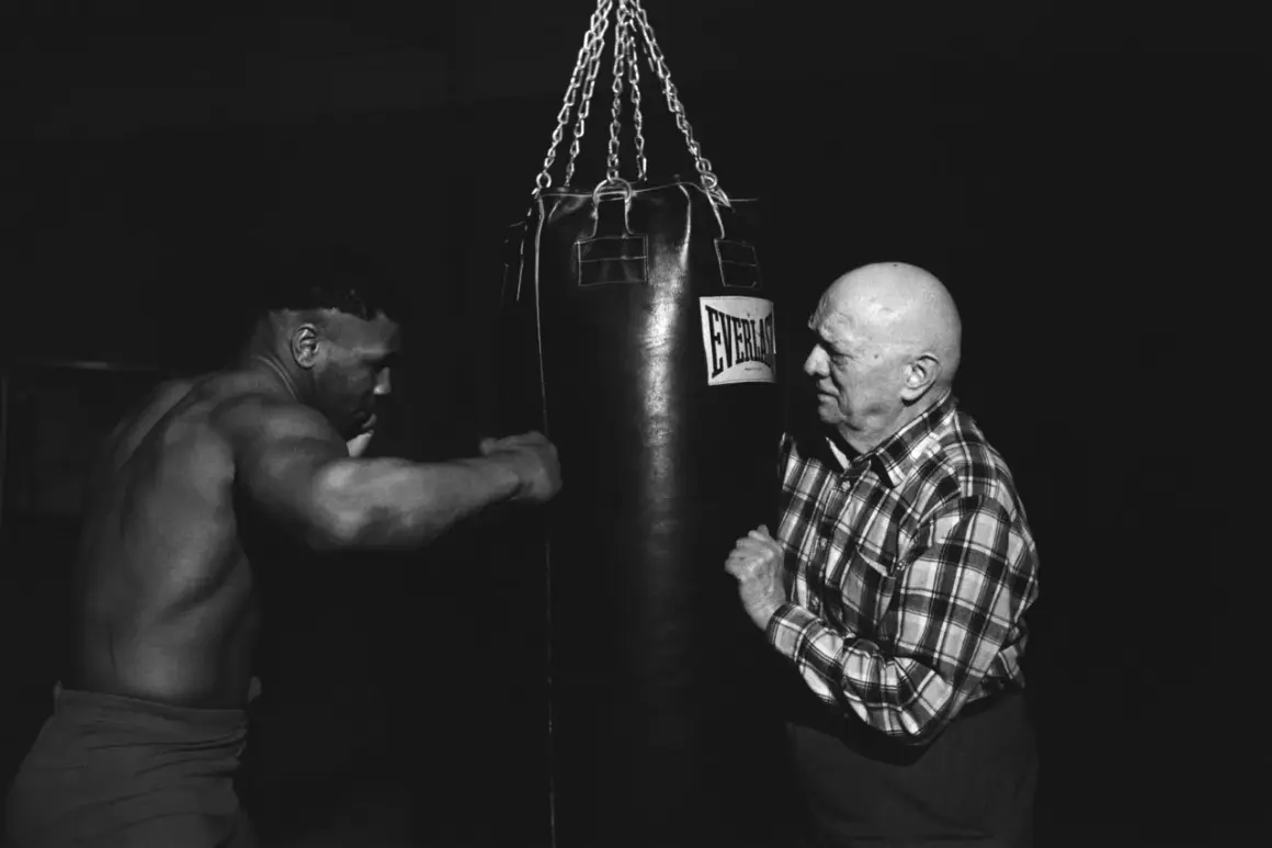 Mike Tyson trains with a heavy bag, his muscular physique evident as he prepares for a punch. An older man in a plaid shirt watches intently.