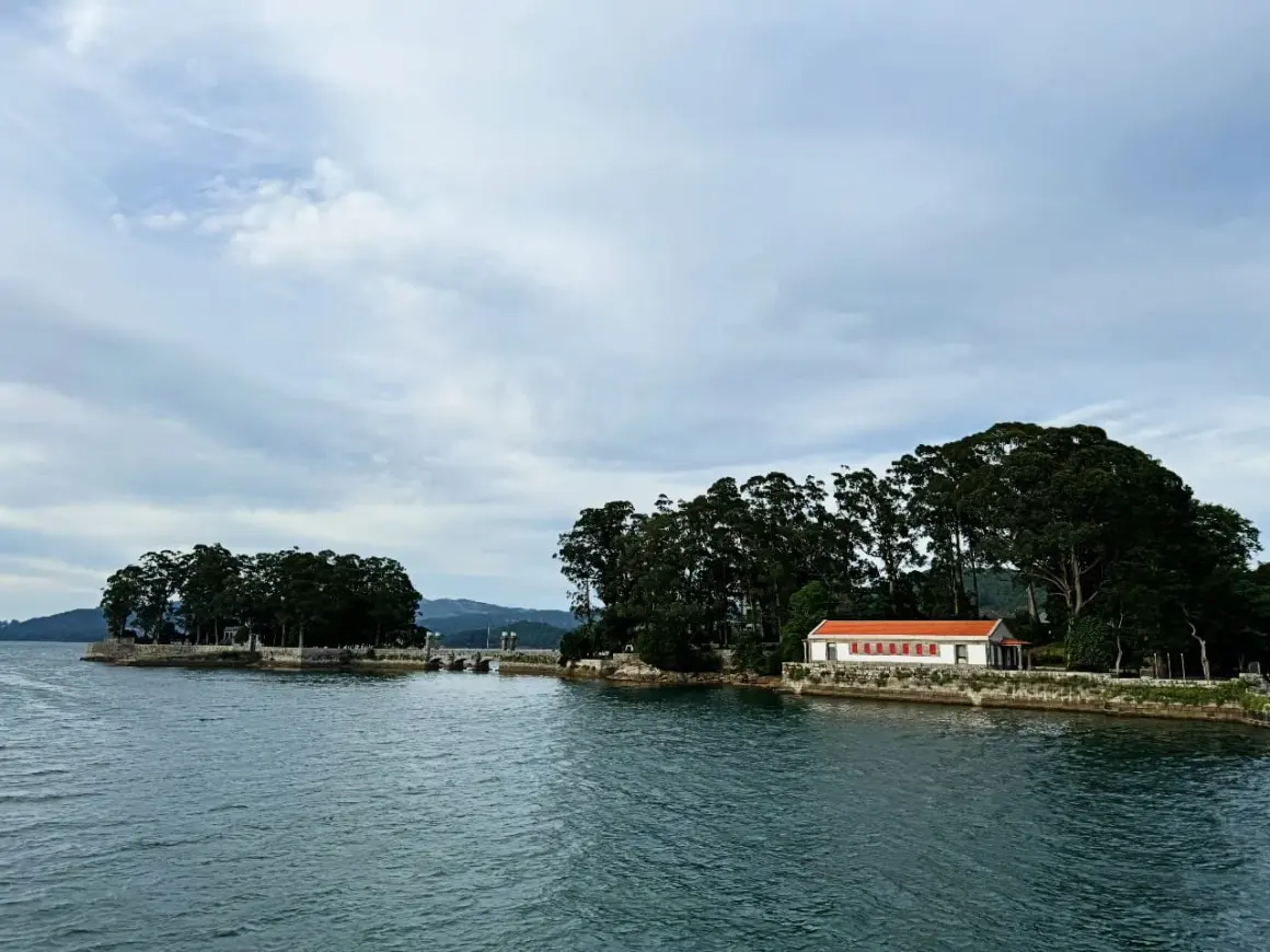 La isla de San Simón, con su casa de tejado naranja y frondosos árboles, se alza sobre el agua serena bajo un cielo nublado.