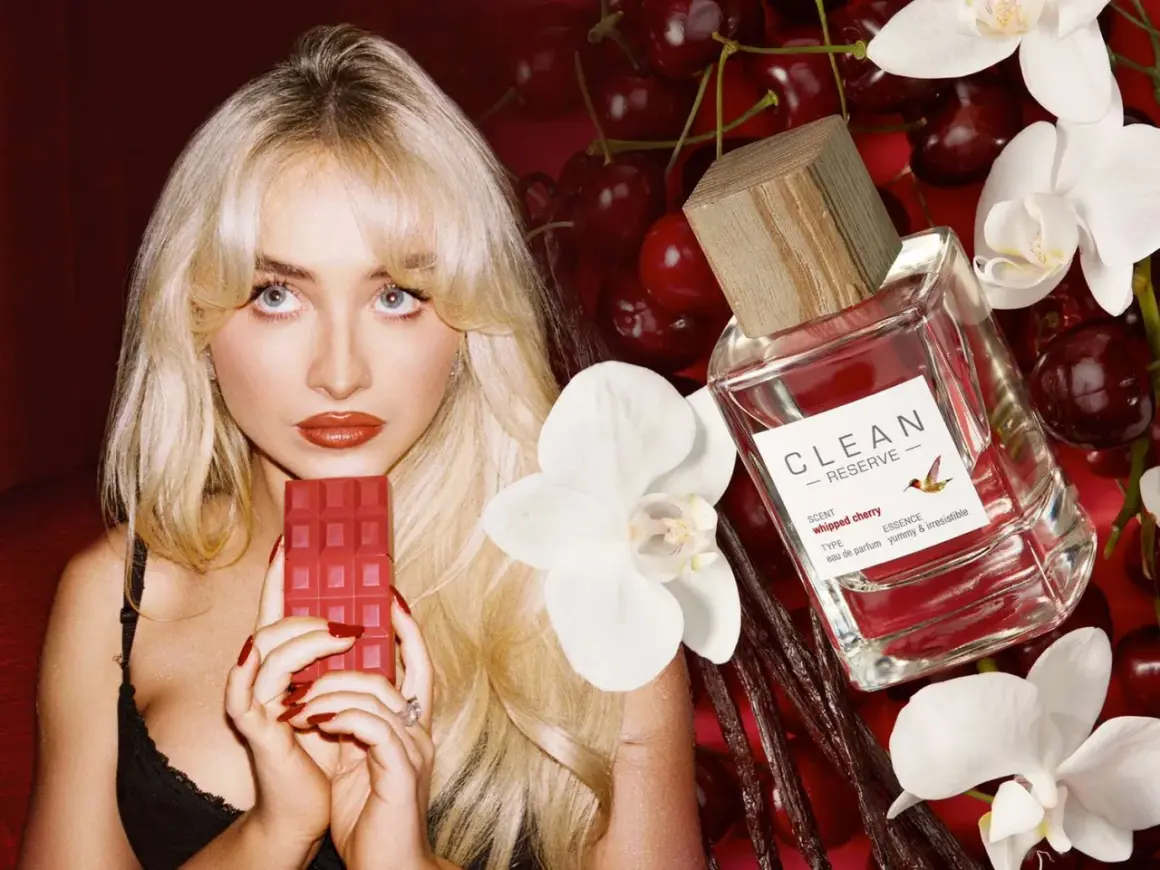 Kobieta z blond włosami trzyma czerwony batonik czekoladowy obok butelki perfum Clean Reserve Whipped Cherry, wiśni i białych kwiatów.