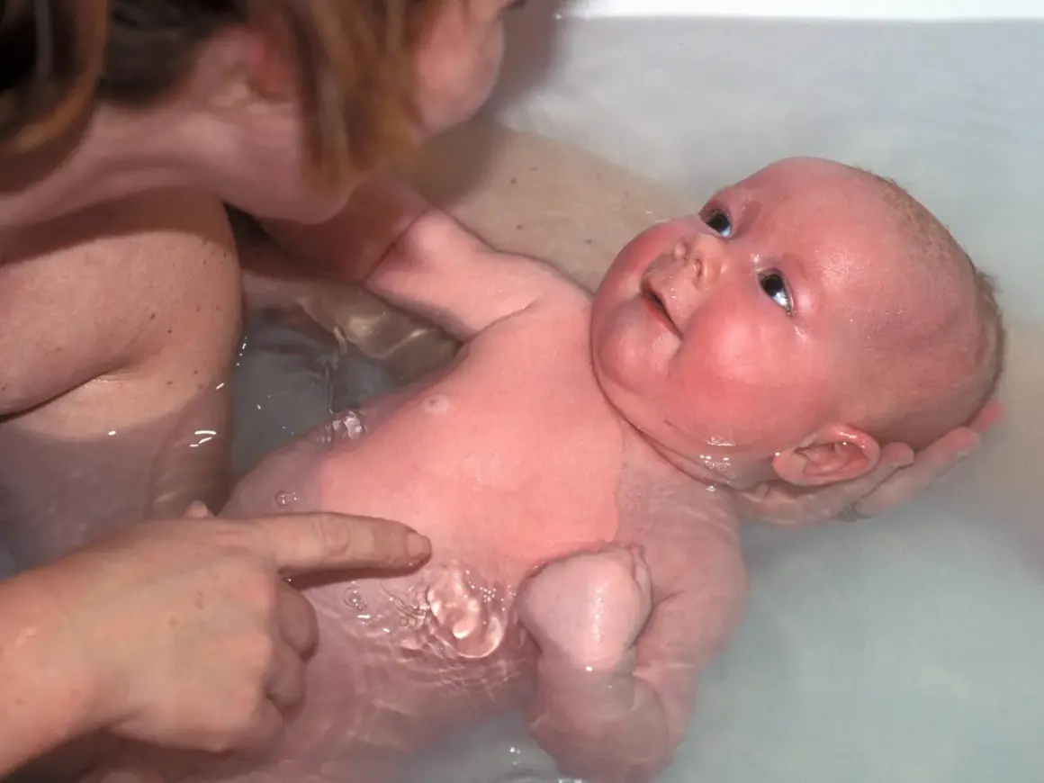Ein Baby genießt sein Bad, während eine Hand es sanft hält und eine andere auf seine Haut zeigt.