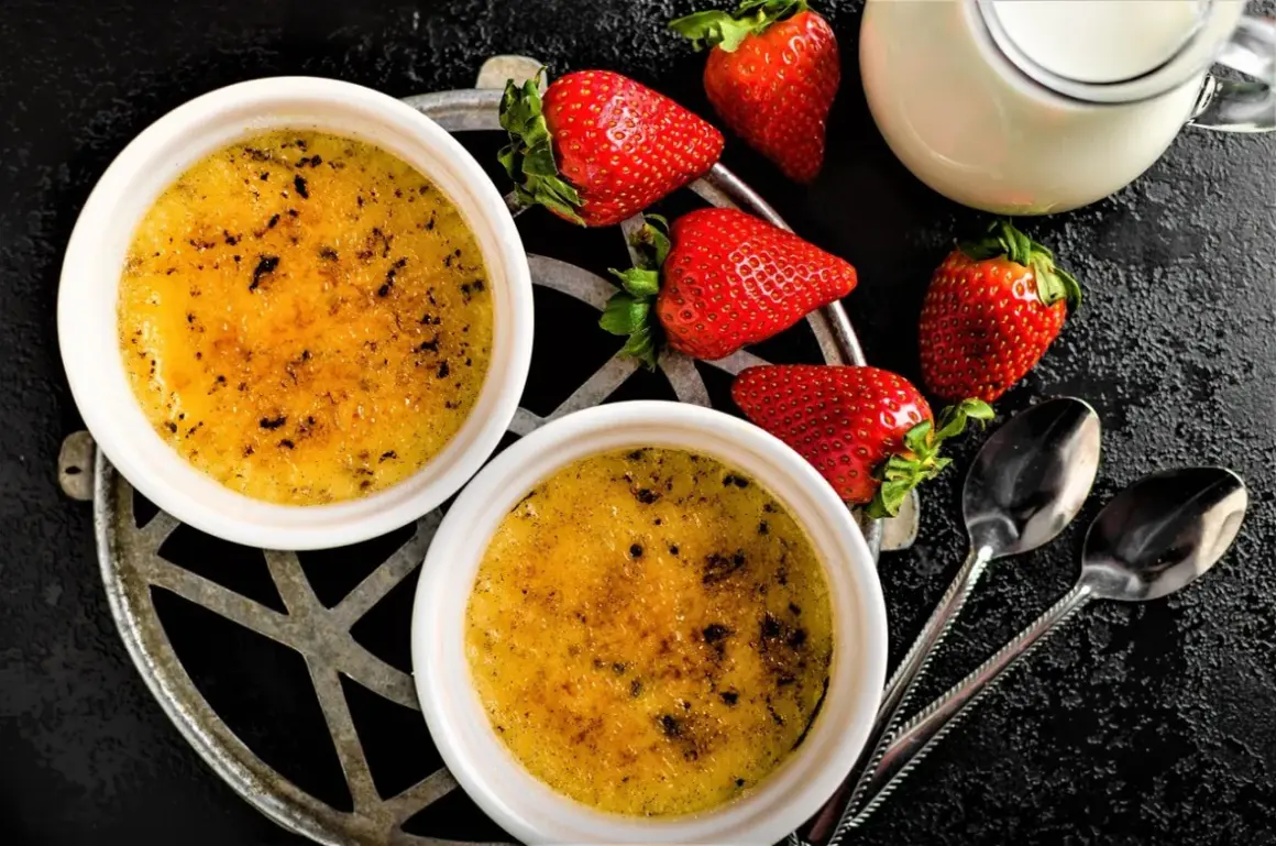Zwei cremige Crème brûlée mit karamellisierter Kruste, garniert mit frischen Erdbeeren und einem Milchkännchen.