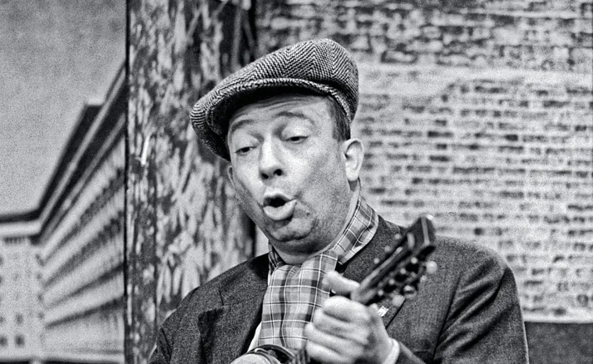 Stanisław Bard Starej Warszawy, w czapce i z gitarą, śpiewa na tle ceglanej ściany.