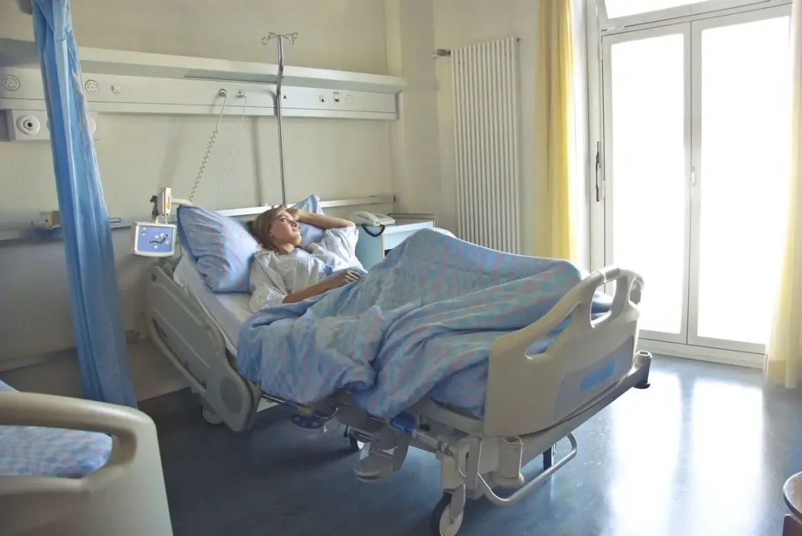 Młoda kobieta leży w szpitalnym łóżku, otulona niebieskim kocem. Jej hospitalizacja przebiega spokojnie, a przez okno wpada jasne światło.