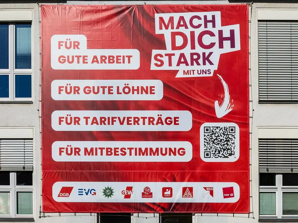 Großes rotes Banner mit der Aufschrift "Mach dich stark mit uns". Es wirbt für gute Arbeit, Löhne, Tarifverträge und Mitbestimmung. Was ist ver.di? Die Antwort steht unten.