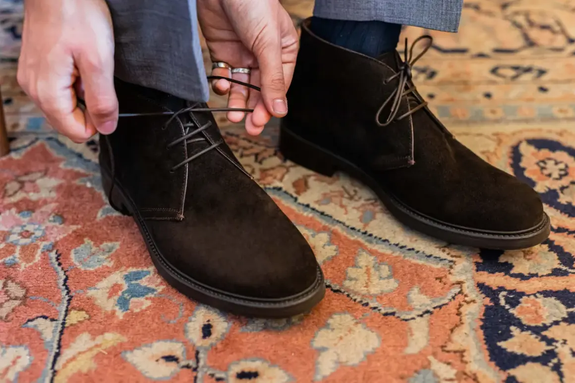 Mani che allacciano stivaletti scamosciati marroni, un dettaglio di stile per polacchi uomo eleganti su un tappeto persiano.