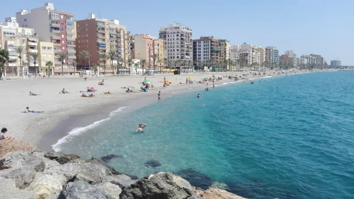 Gente disfrutando del sol y el mar en la playa del Zapillo, con edificios residenciales al fondo.