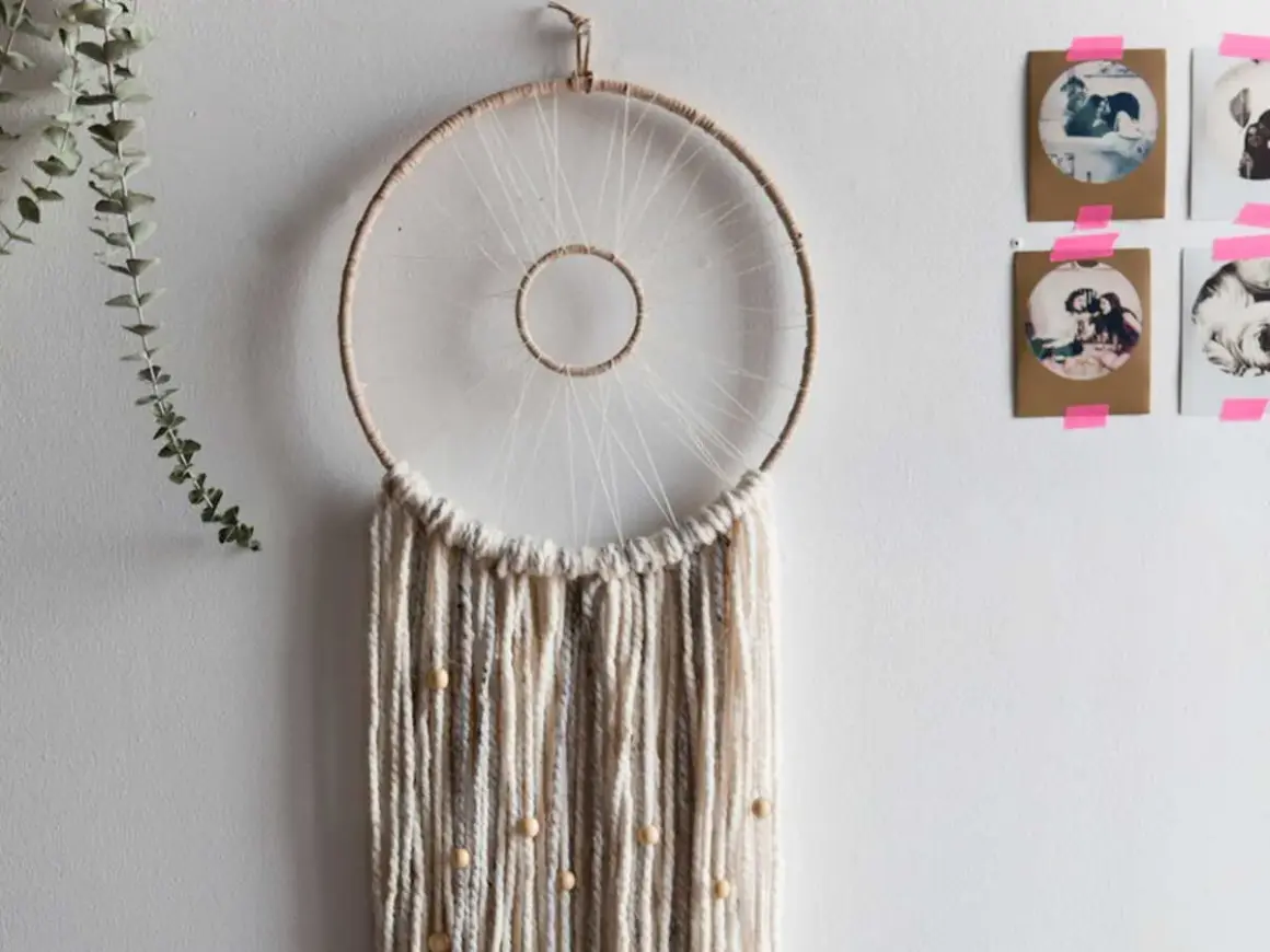 Un attrape-rêves fait main, parfait pour un tuto attrape rêve. Des fils de laine beiges et gris tombent, ornés de perles en bois.