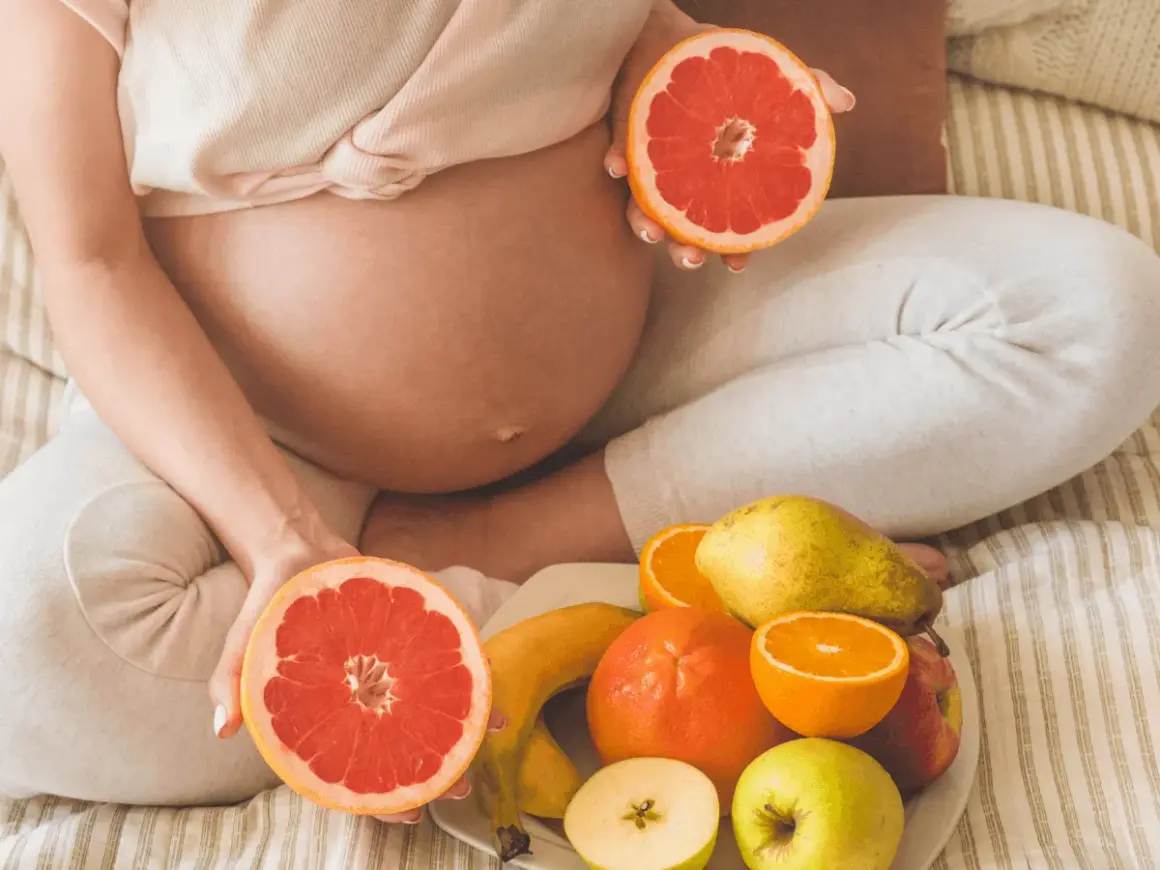 Ciężarna kobieta trzyma połówki grejpfruta, obok talerz z owocami. Czy w ciąży można jeść pomarańcze? Tak, to zdrowe!