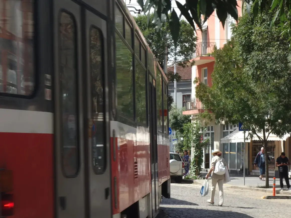 Przejeżdżający tramwaj w słoneczny dzień w Sofii. Kobieta w kapeluszu idzie ulicą z torbami.