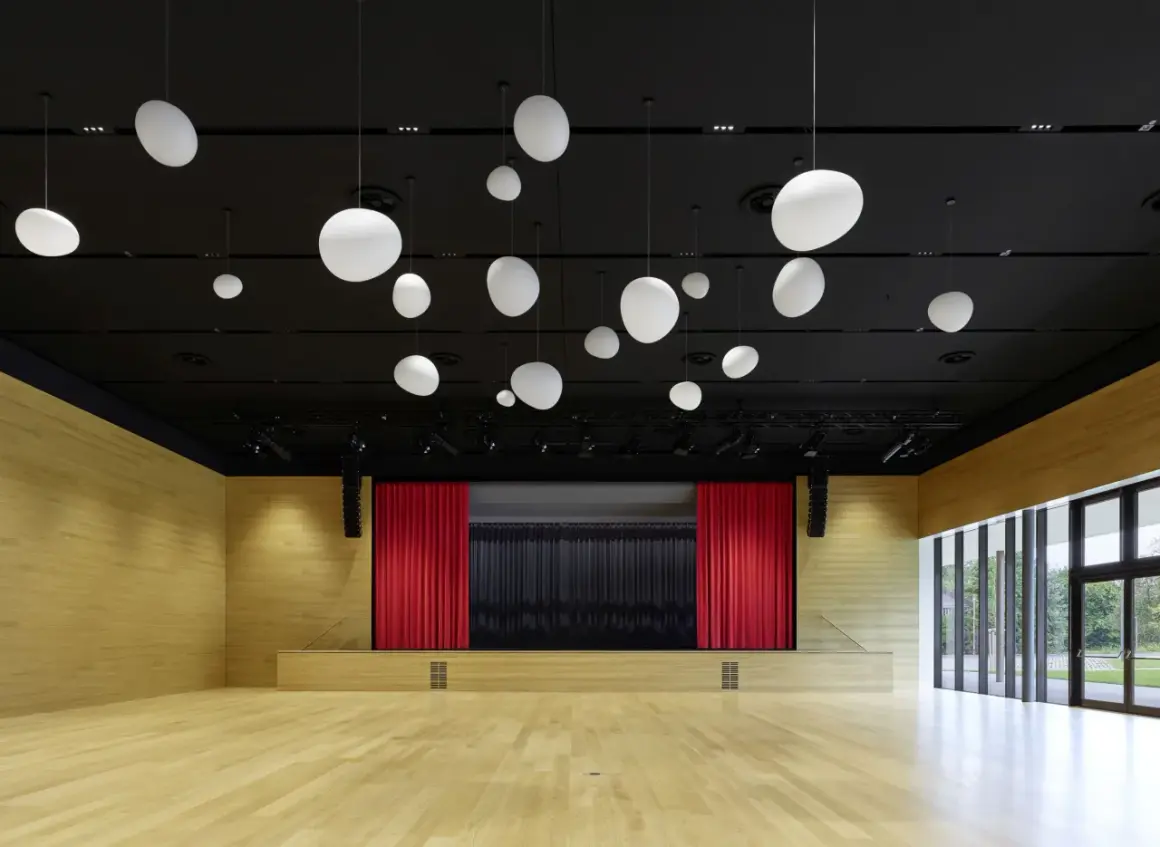 Moderne Aula mit Holzwänden, Bühne mit roten Vorhängen und vielen weißen Hängelampen. Die nobelgusch pfedelbach ist bereit für Veranstaltungen.