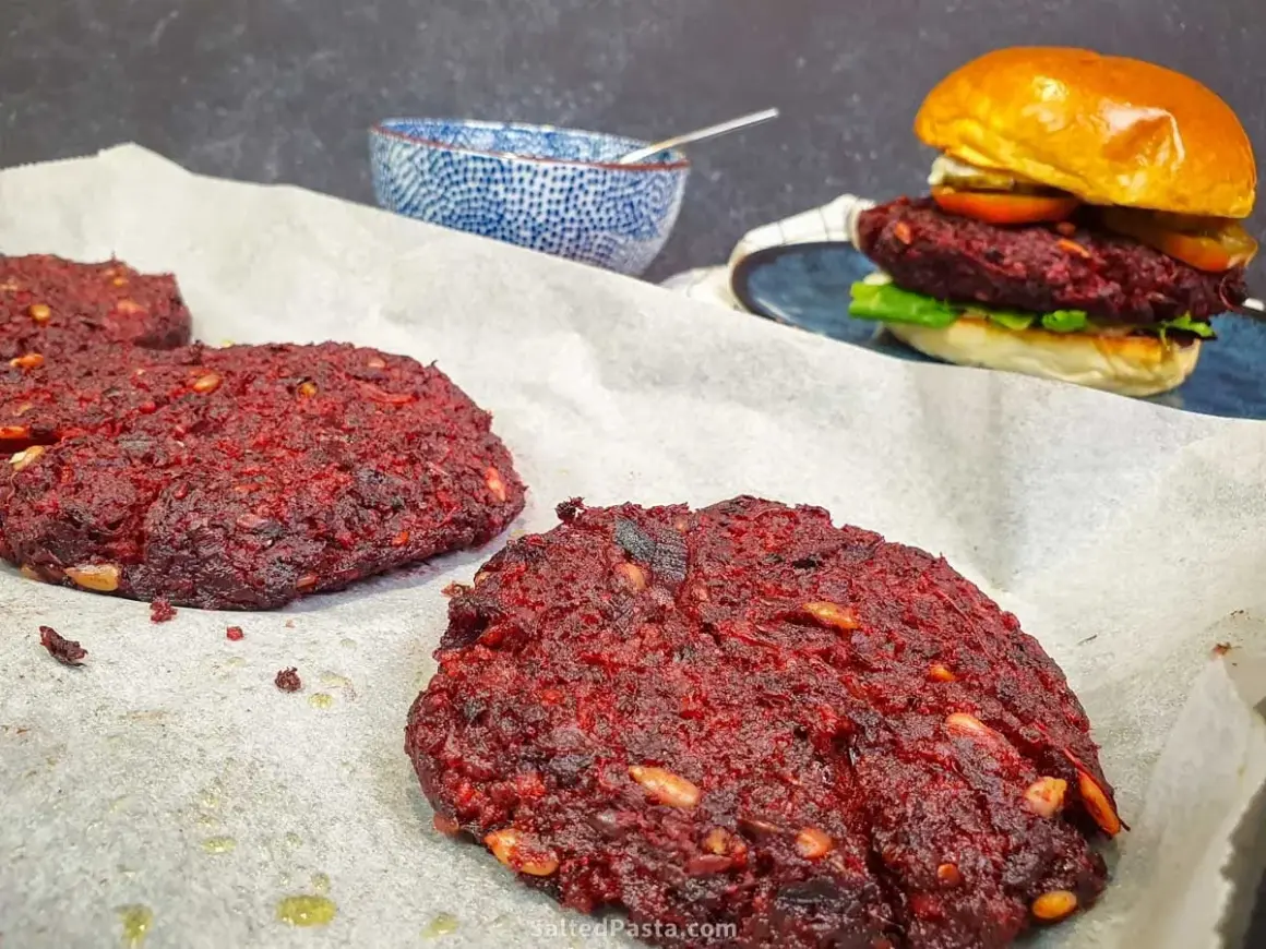 Gotowe burgery z buraka na papierze do pieczenia, jeden złożony burger z bułką i dodatkami w tle.