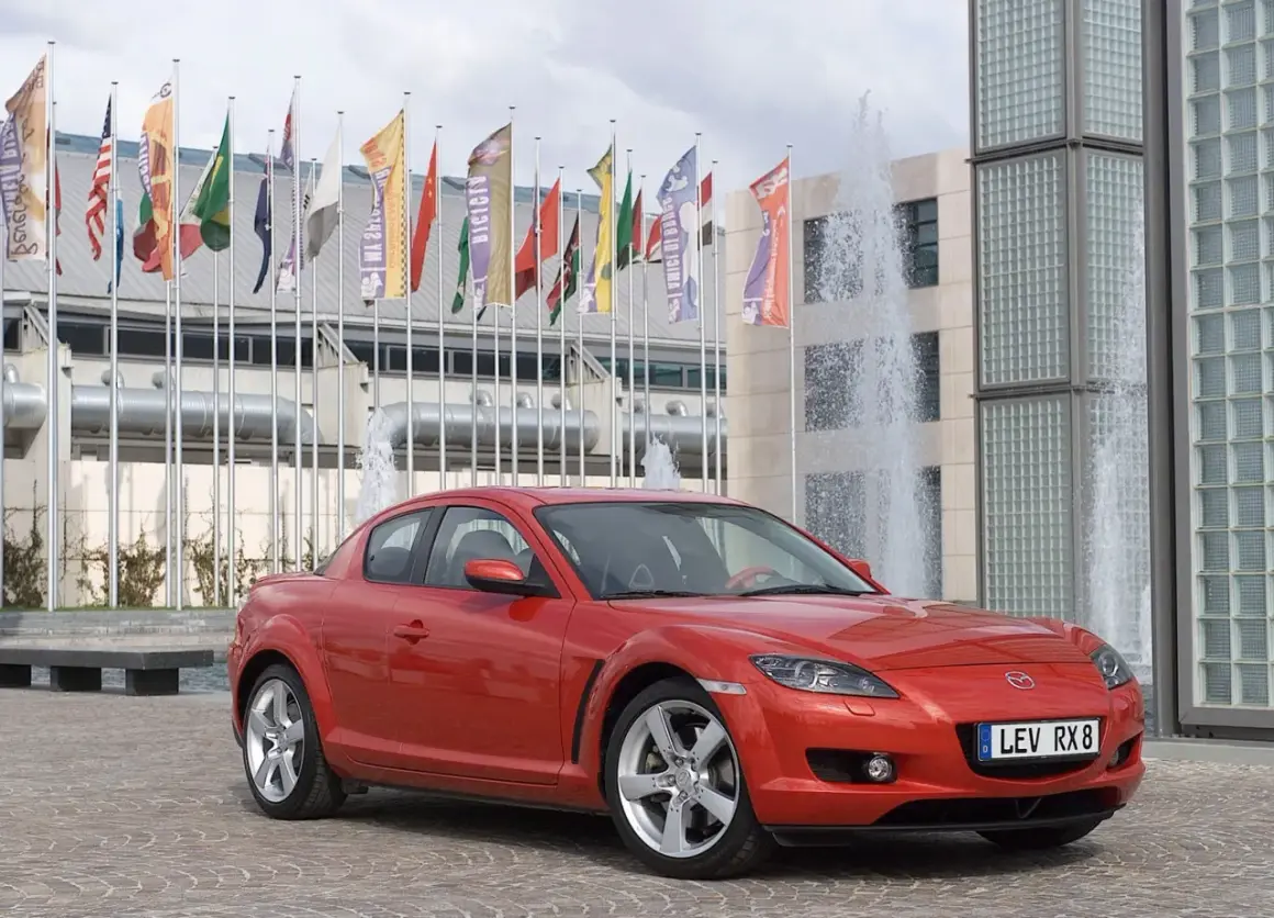Czerwona Mazda RX-8 zaparkowana przed budynkiem z fontannami i flagami.