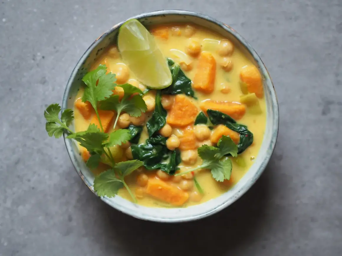 Aksamitne curry z ciecierzycy z kawałkami batata i szpinaku, ozdobione świeżą kolendrą i ćwiartką limonki.