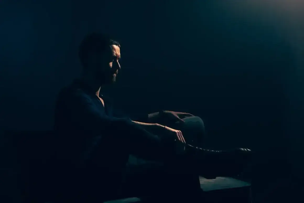 Mężczyzna z brodą siedzi w półmroku, wpatrzony w dal. Jego postawa jest **low key**, emanuje spokojem i zadumą.
