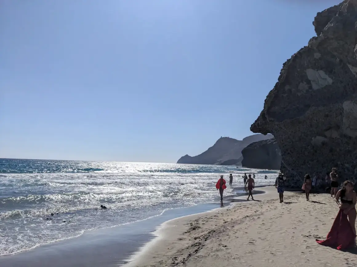 Gente pasea por la **playa de Mónsul** bajo un cielo azul, con olas rompiendo en la orilla y una gran roca a la derecha.