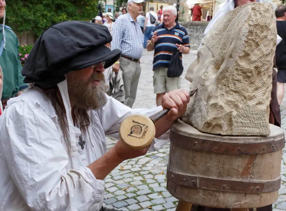 Steinmetz bei der Arbeit am Bad Wimpfen Zunftmarkt. Ein Mann mit Bart und schwarzer Kappe meißelt an einer Steinskulptur.