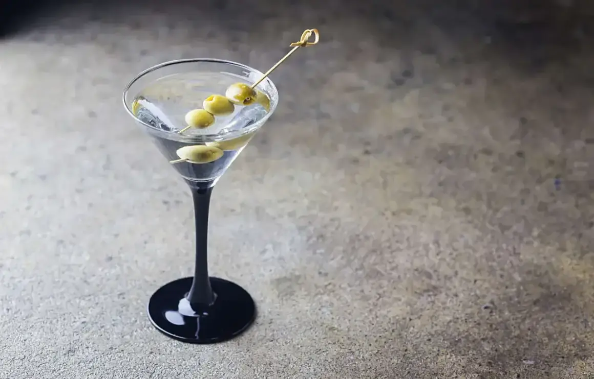 Elegancki dirty martini z trzema oliwkami na wykałaczce, stojący na czarnej podstawie.