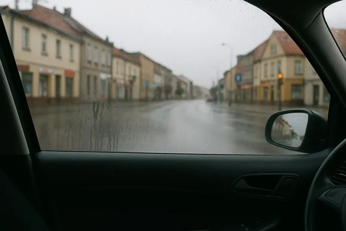 Widok z wnętrza samochodu na deszczową ulicę. Krople na szybie i wilgoć w samochodzie utrudniają widoczność.