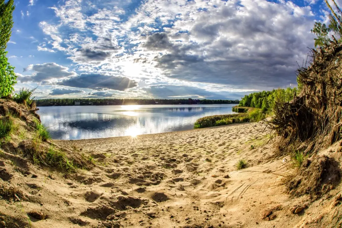 Piaskowa plaża nad jeziorem, słońce przebija się przez chmury, tworząc refleksy na wodzie. W oddali las, a po prawej stronie widoczne korzenie drzew. Idealne miejsce na wypoczynek nad pogorią zalew.