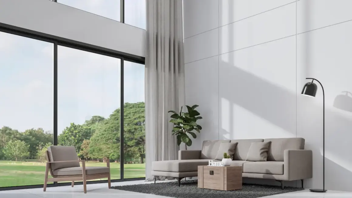 Nowoczesny salon z wysokim sufitem, szara sofa, fotel i wielkie okna z widokiem na zieleń.