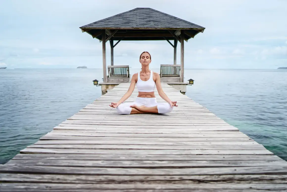 Frau in weißer Kleidung meditiert auf einem Steg am Meer. Eine Anleitung zur transzendentalen Meditation, um inneren Frieden zu finden.