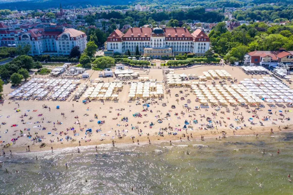 Widok z lotu ptaka na zatłoczoną plażę z białymi parasolami i hotelami w tle.