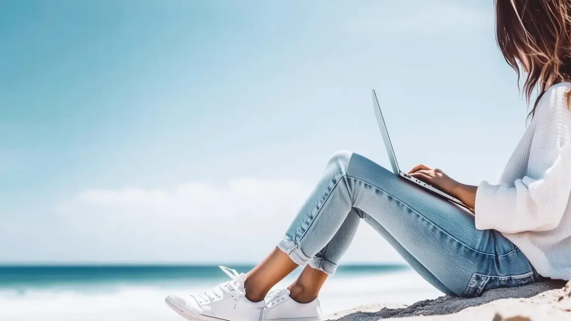 Junge Frau mit Laptop am Strand, genießt die Sonne und das Meer. Ein perfekter Ort für ihr IU Erasmus-Projekt.
