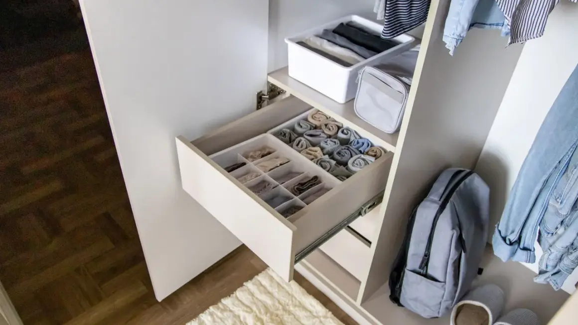 Schrank mit ordentlich gefalteten Kleidungsstücken in Schubladen und Körben. Ein Rucksack und Jeans hängen daneben. Zu viele Klamotten zu wenig Platz? Hier ist die Lösung!
