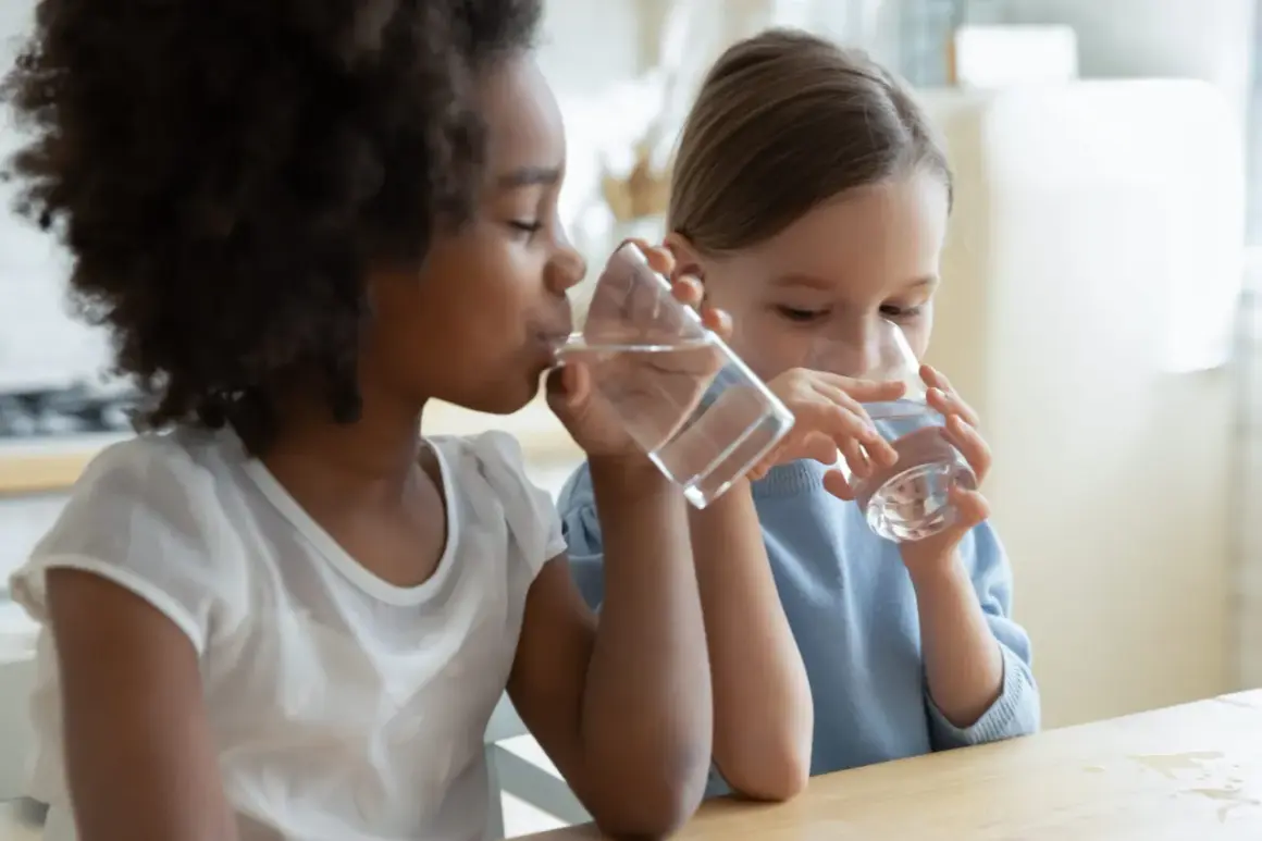 Deux enfants, une fille noire et une fille blanche, boivent de l'eau à la même table. L'eau est essentielle pour chaque enfant.