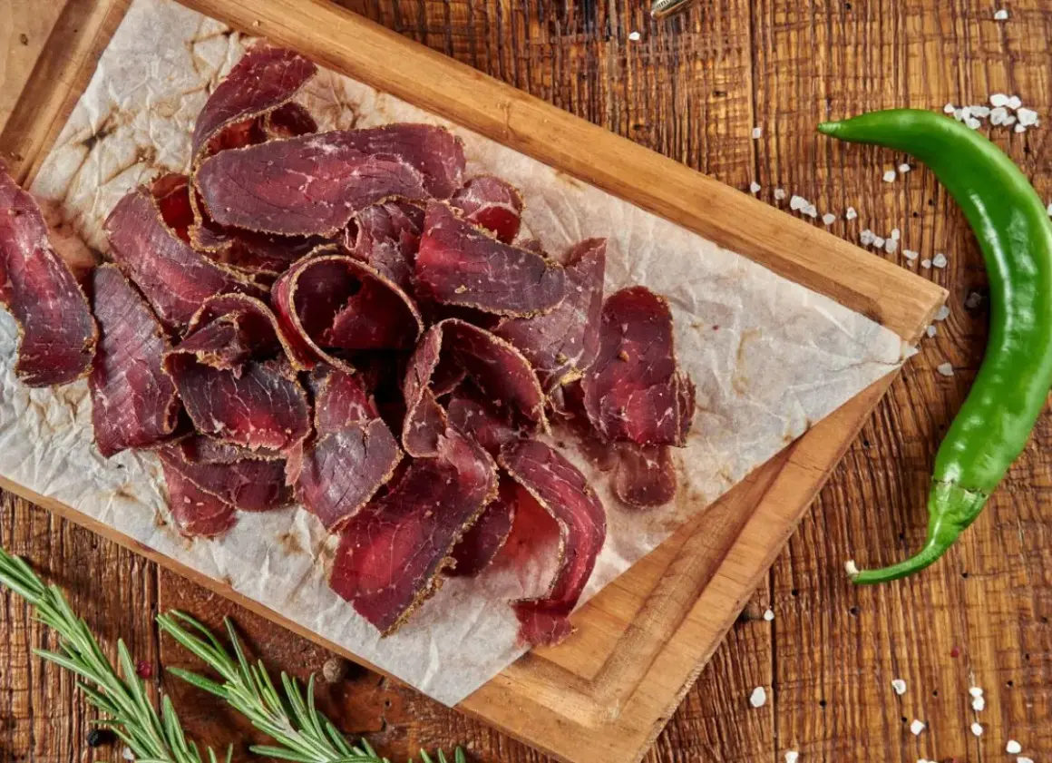 Suszona wołowina przepis na pyszne przekąski. Na desce ułożone cienkie plastry mięsa, obok zielona papryczka chili i rozmaryn.