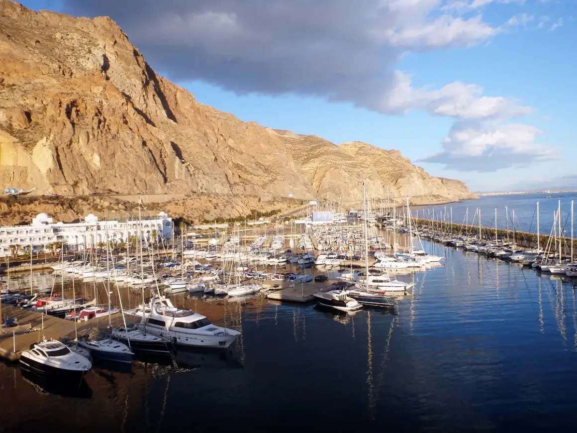 El puerto deportivo Aguadulce, con sus barcos amarrados y un hotel blanco, se extiende bajo una imponente montaña rocosa.