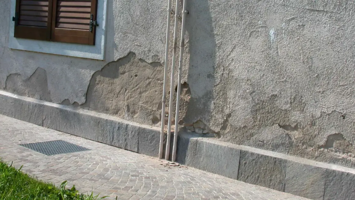 Il muro esterno grezzo mostra scrostamenti e necessita di essere pitturato. Tre tubi bianchi corrono verticalmente lungo la parete.