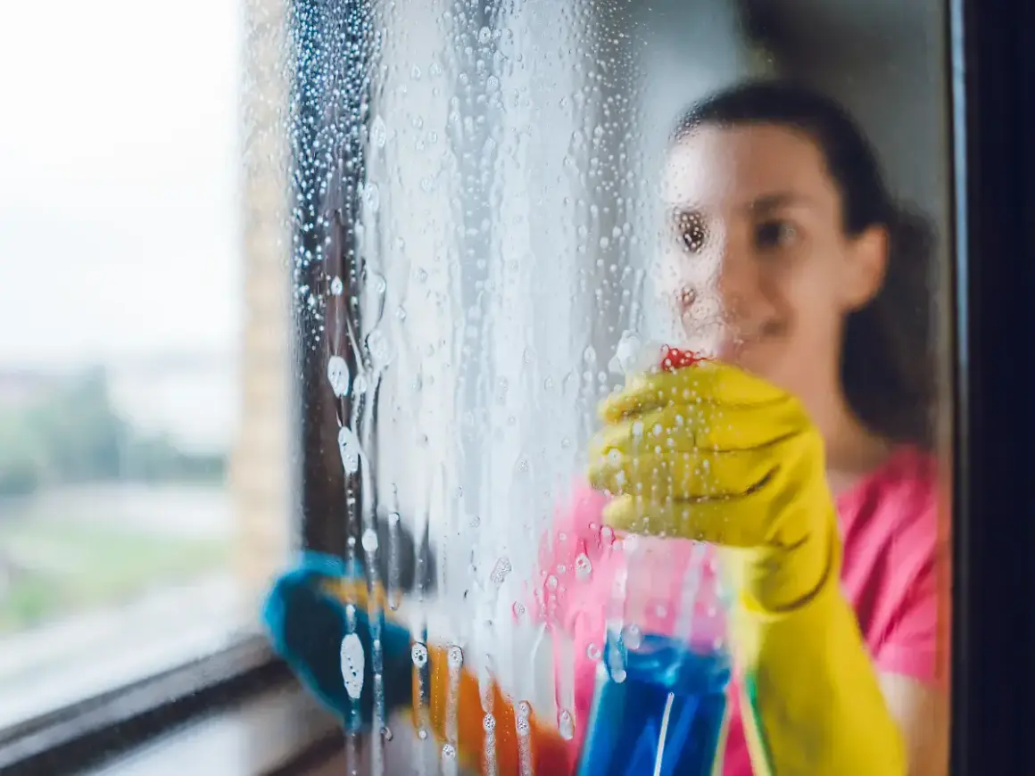 Frau mit gelben Handschuhen sprüht Reinigungsmittel auf ein Fenster, um es zu putzen.