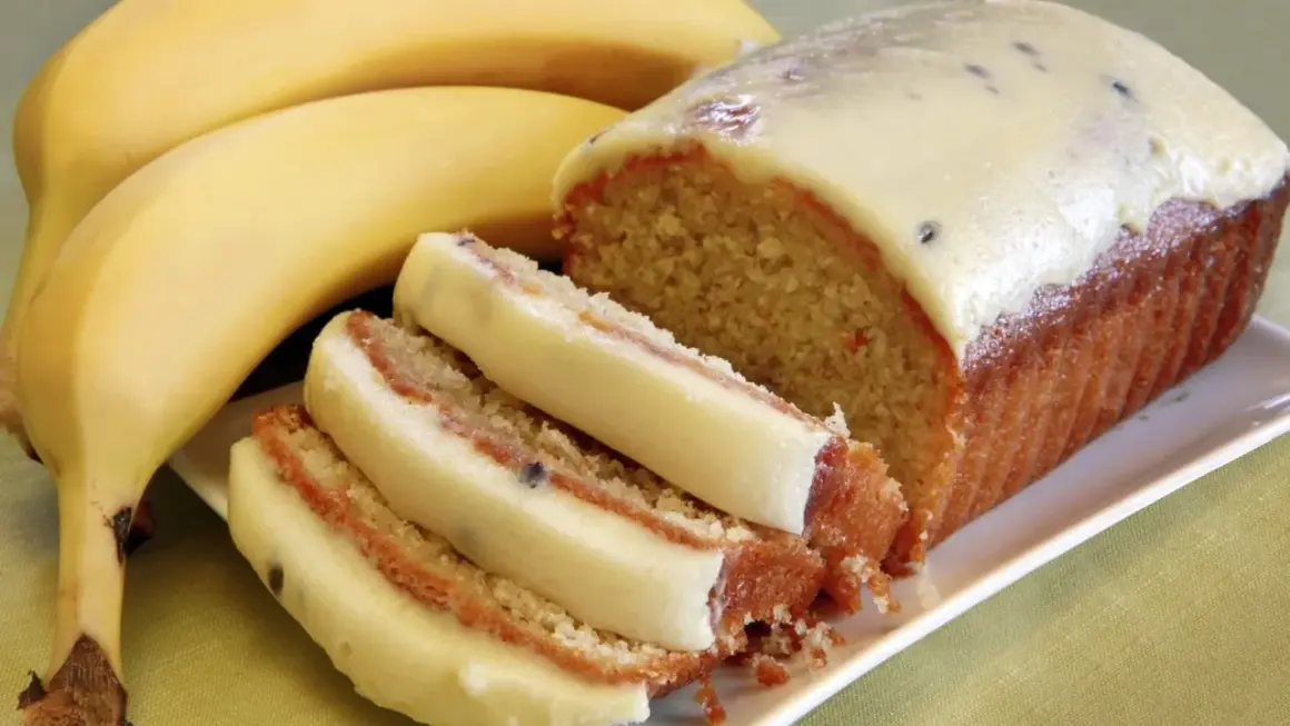 Wilgotne ciasto bananowe z kremową polewą i kawałkami owoców, obok dojrzałych bananów.
