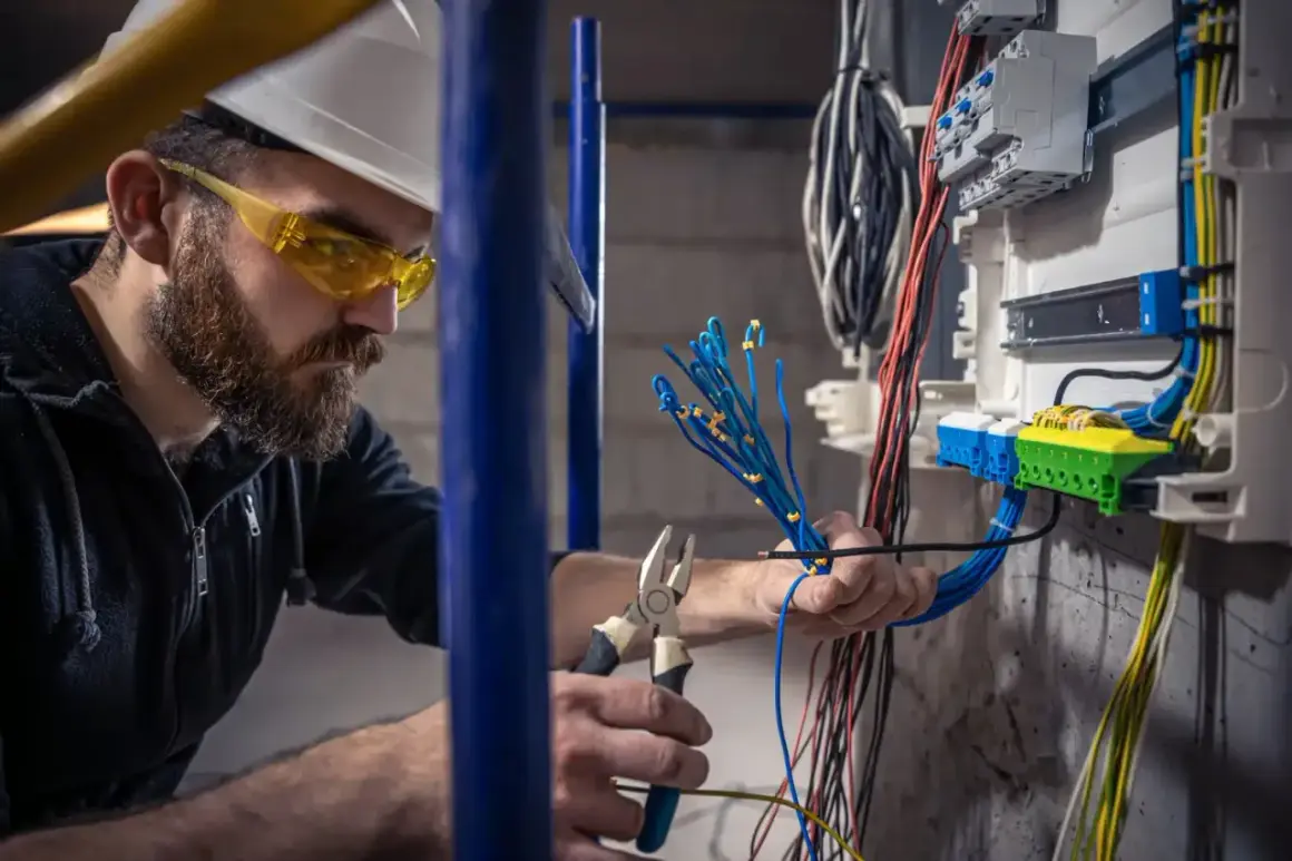 Elektryk w okularach ochronnych i kasku pracuje przy skrzynce z przewodami. Sprawdza, co daje sep g1, podłączając niebieskie kable.