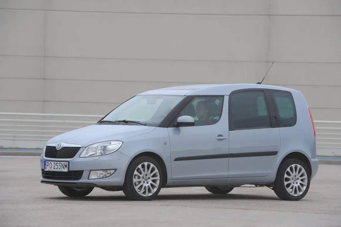 Jasnoszary Skoda Roomster z kierowcą za kierownicą stoi na betonowym placu przed szarym budynkiem.