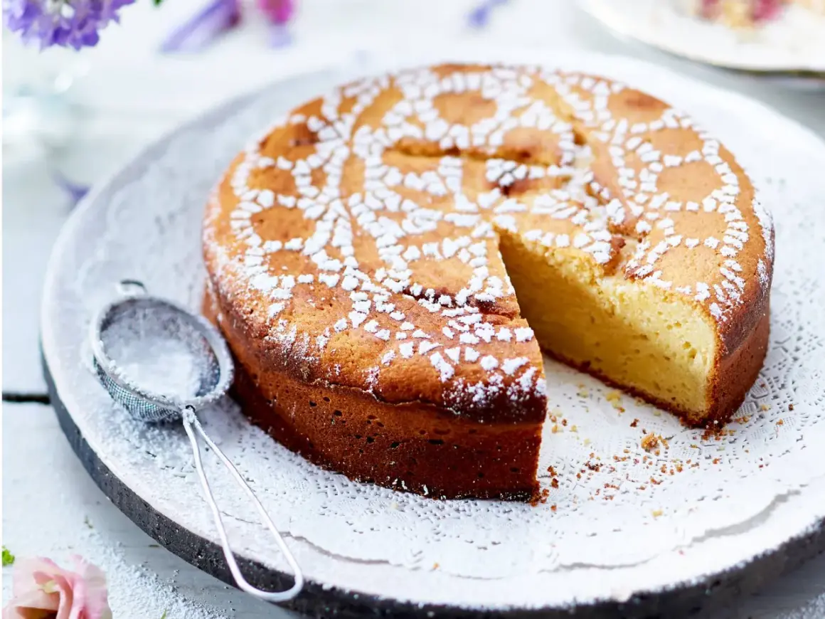 Ein saftiger Joghurt Kuchen, mit Puderzucker bestäubt und einem Stück herausgeschnitten. Daneben liegt ein kleines Sieb.