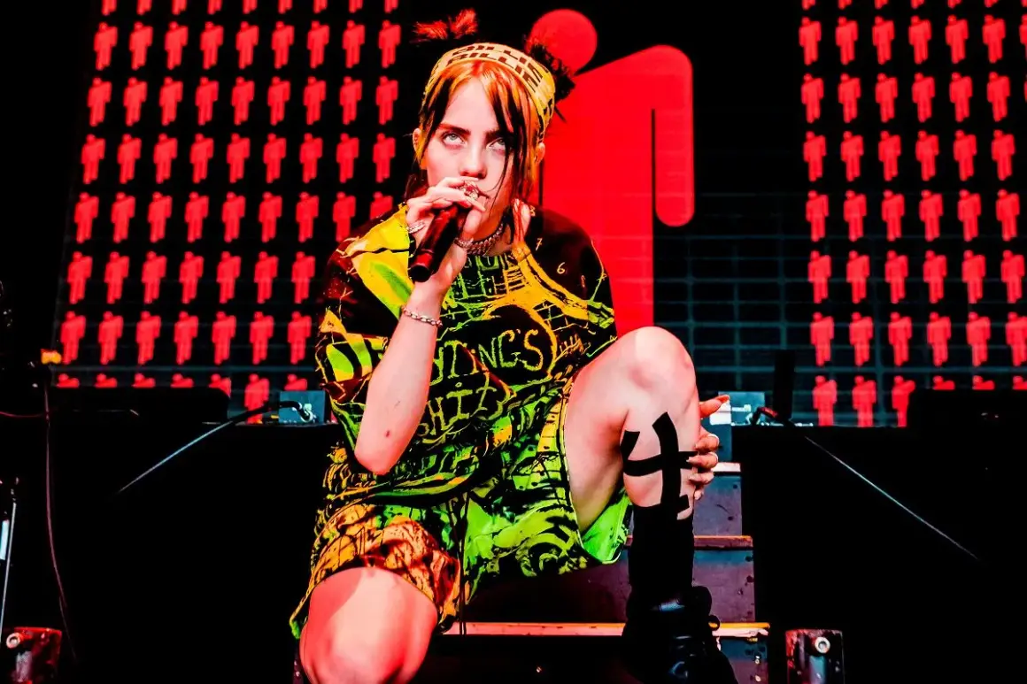 Billie Eilish na koncercie, w neonowym stroju, z mikrofonem w dłoni. Tło zdobią czerwone sylwetki ludzi.