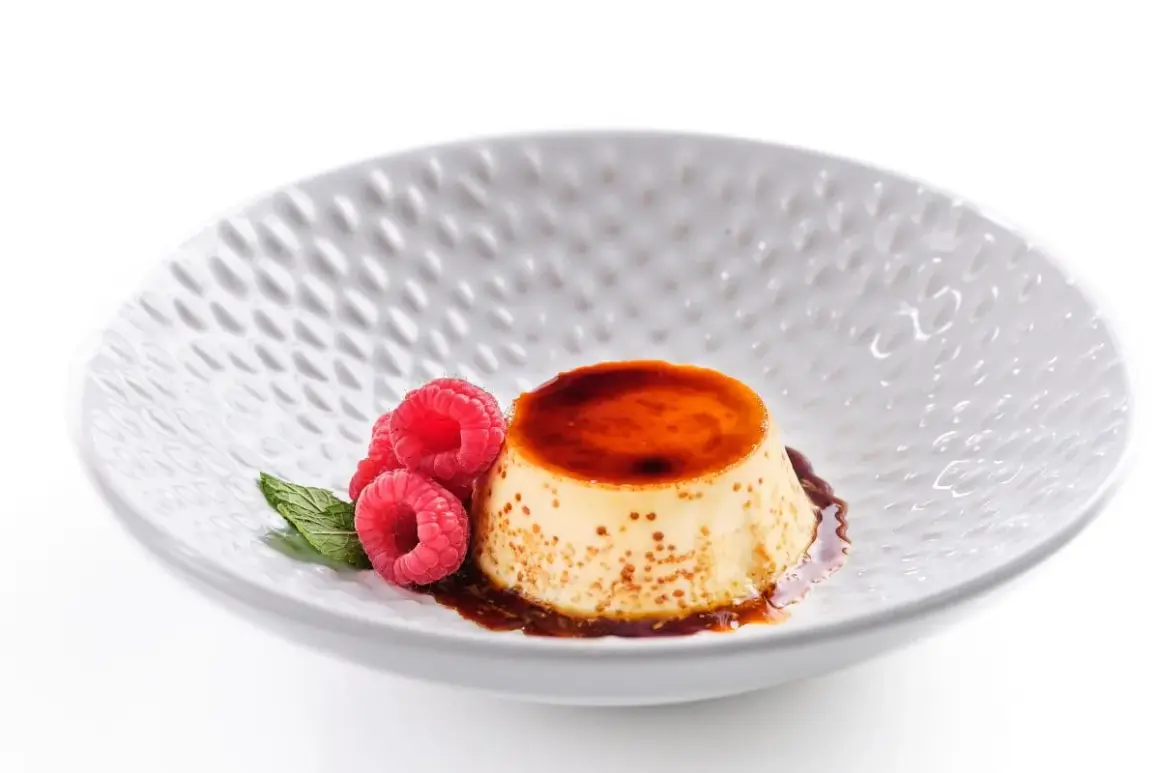 Delicioso flan de queso fresco con caramelo líquido, decorado con frambuesas y menta en un plato blanco texturizado.