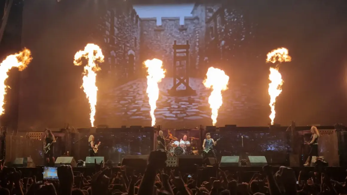 Płomienie buchają na scenie, gdzie grają zespoły rockowe. W tle widać zamek i gilotynę.