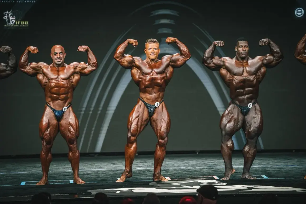 Drei Bodybuilder präsentieren ihre Muskeln auf der Bühne. Man fragt sich, wann ist Mr. Olympia?