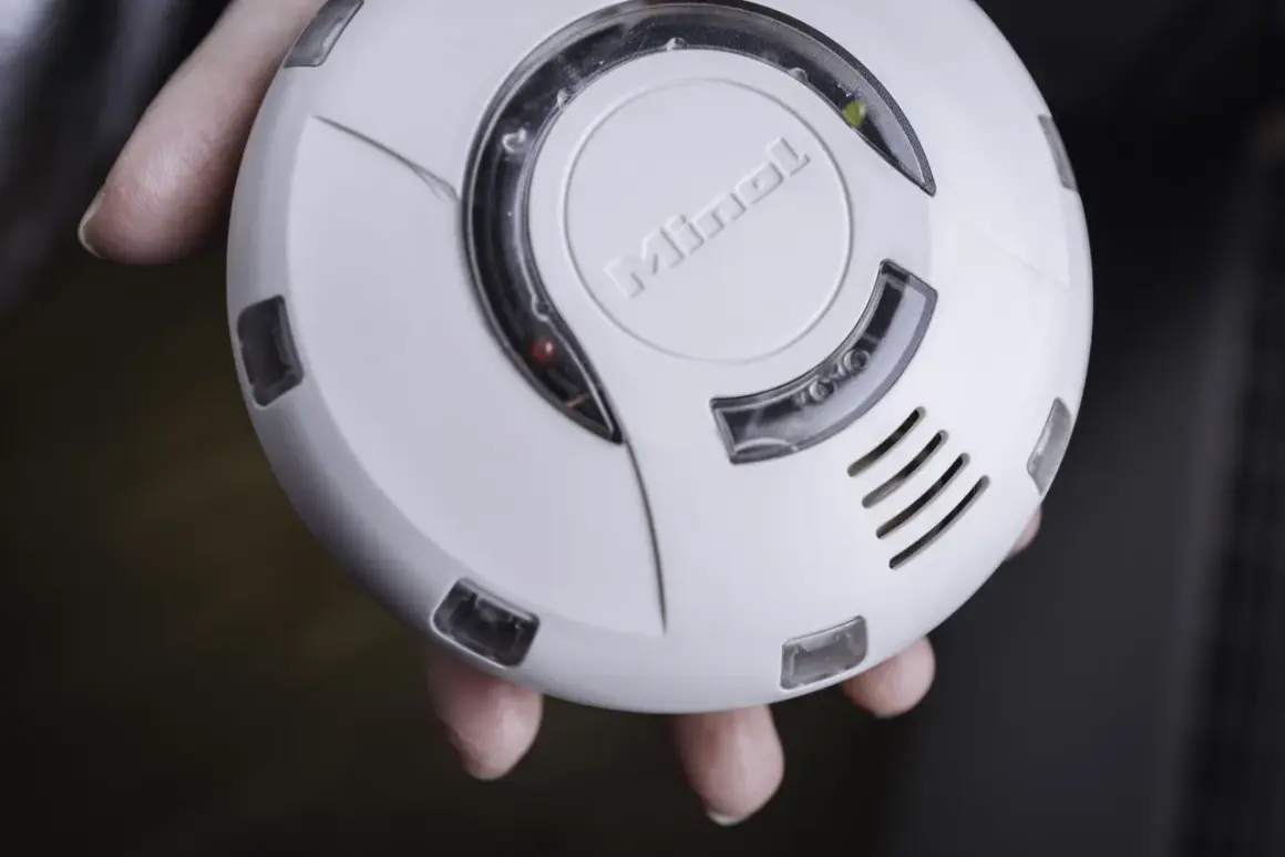 Ein weißer enocean rauchmelder mit dem Schriftzug "Minol" wird von Fingern gehalten. Ein rotes und ein gelbes Licht leuchten.